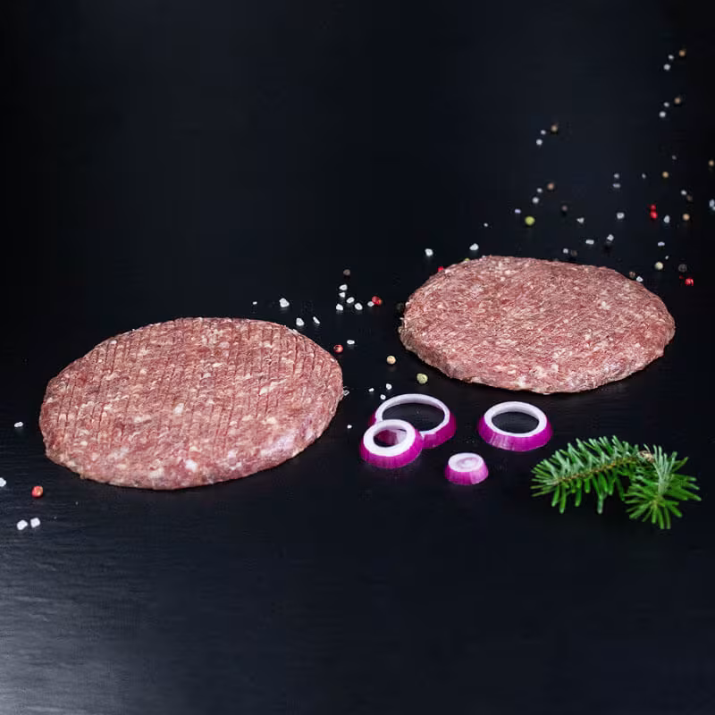 Rohe Wildburger-Patties mit Zwiebelringen, Kräutern und Gewürzen auf dunklem Untergrund