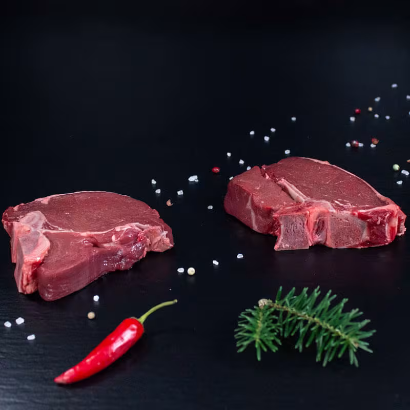 Rohe Hirsch-T-Bone-Steaks mit Kräutern und Gewürzen auf dunklem Untergrund