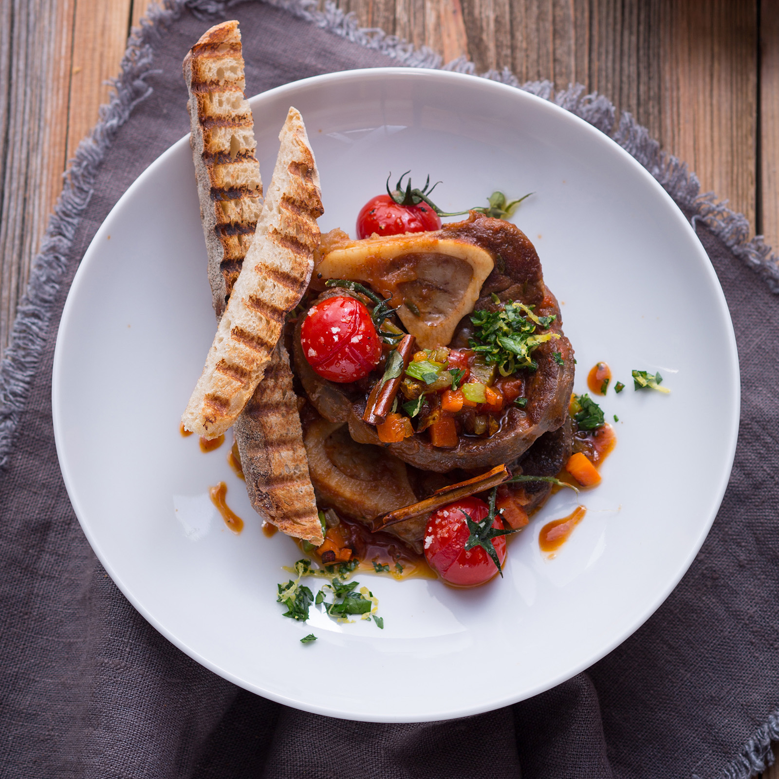 Hirsch-Ossobuco mit Gremolata, geschmorten Tomaten und geröstetem Weißbrot auf weißem Teller