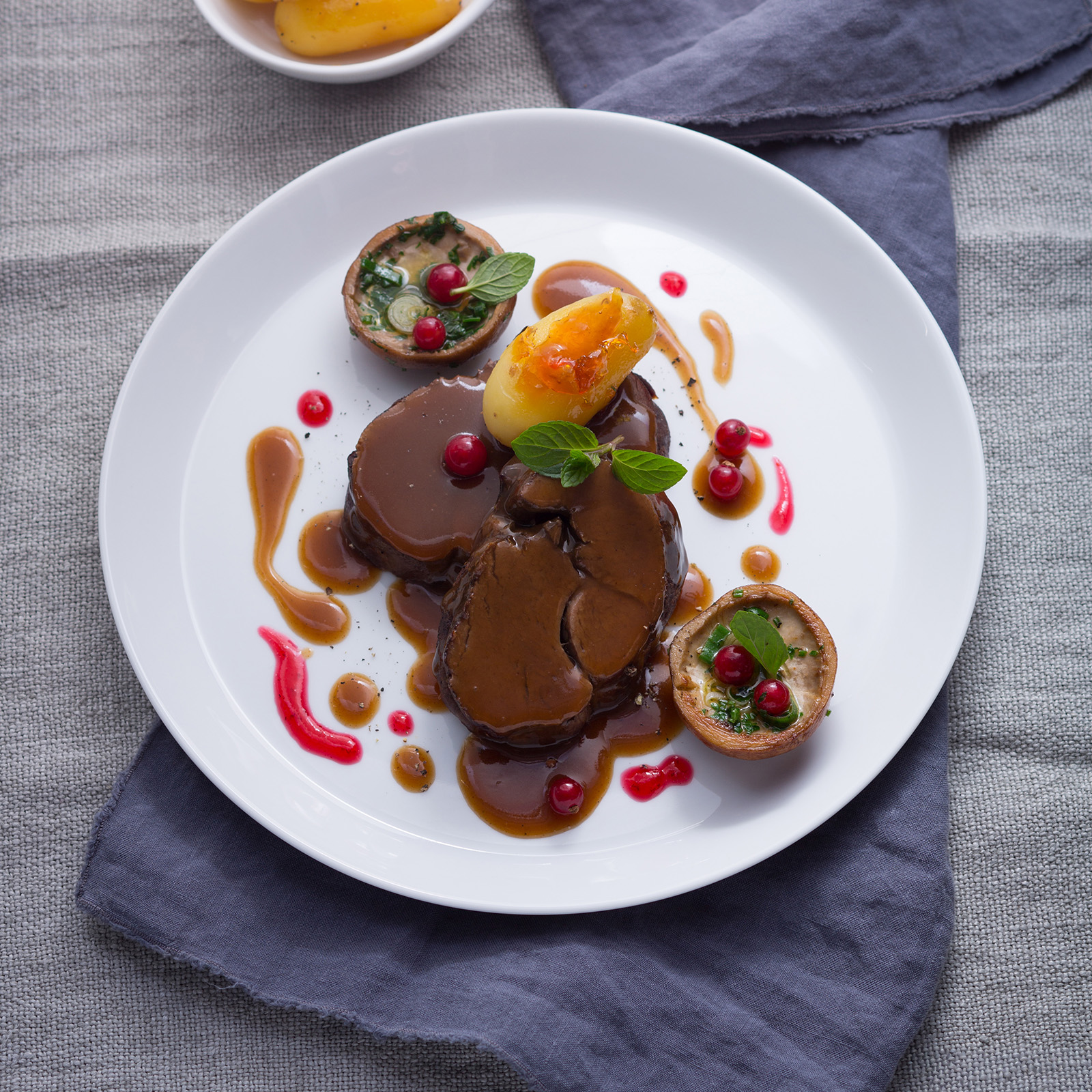 Aufgeschnittener Rehbraten mit dunkler Johannisbeer-Minze-Sauce, roten Johannisbeeren, frischer Minze und gefüllten Champignons
