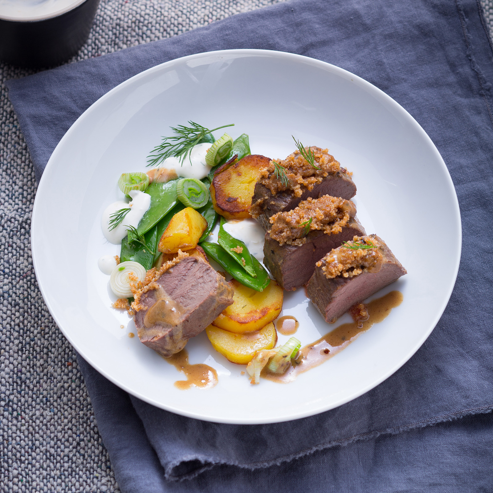 Rehrücken mit Senfkruste, Kartoffeln und Dill-Honig-Sauce auf weißem Teller angerichtet