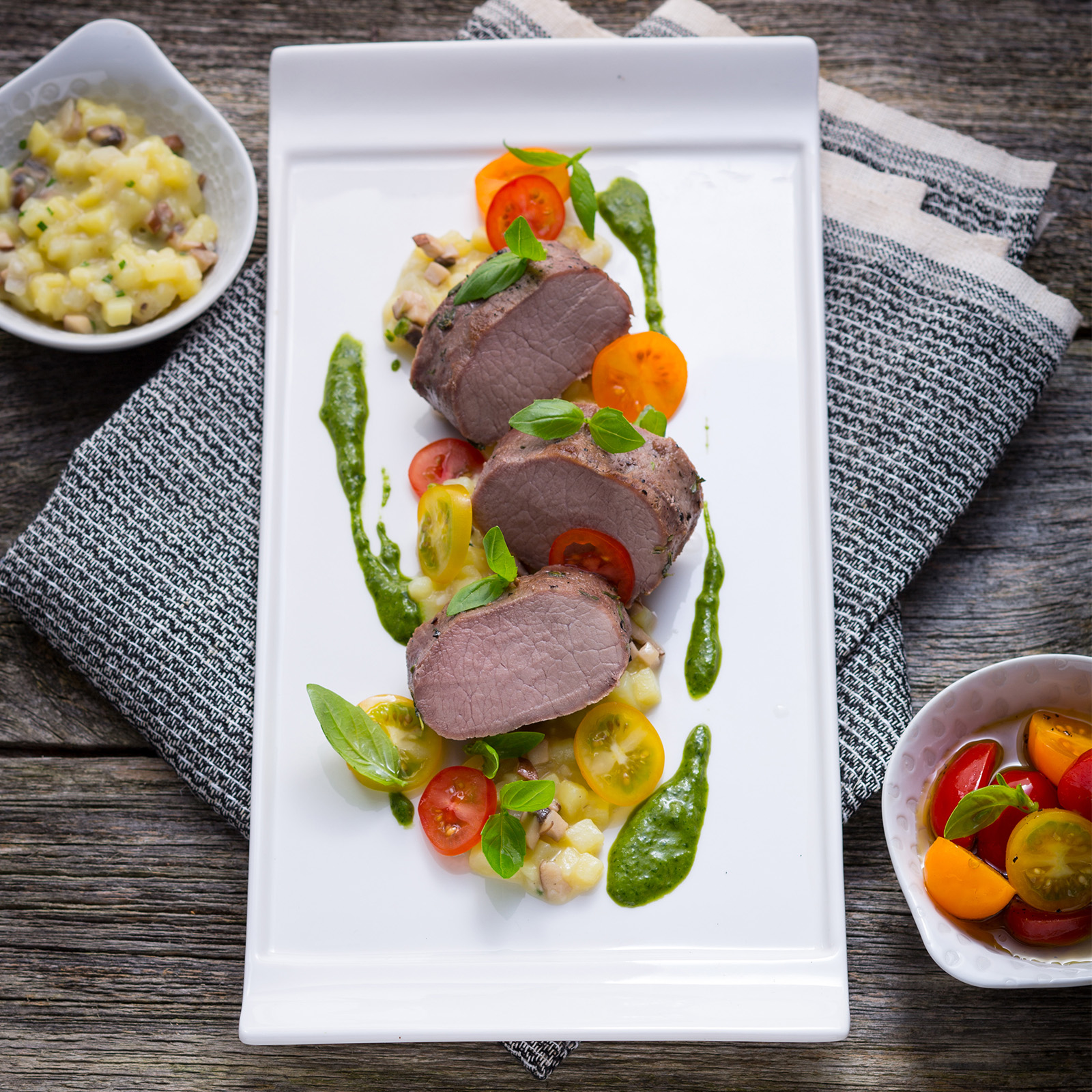 Aufgeschnittener Wildschwein-Frischlingsrücken mit Kräuteröl, bunten Kirschtomaten, Basilikum und Kartoffelsalat