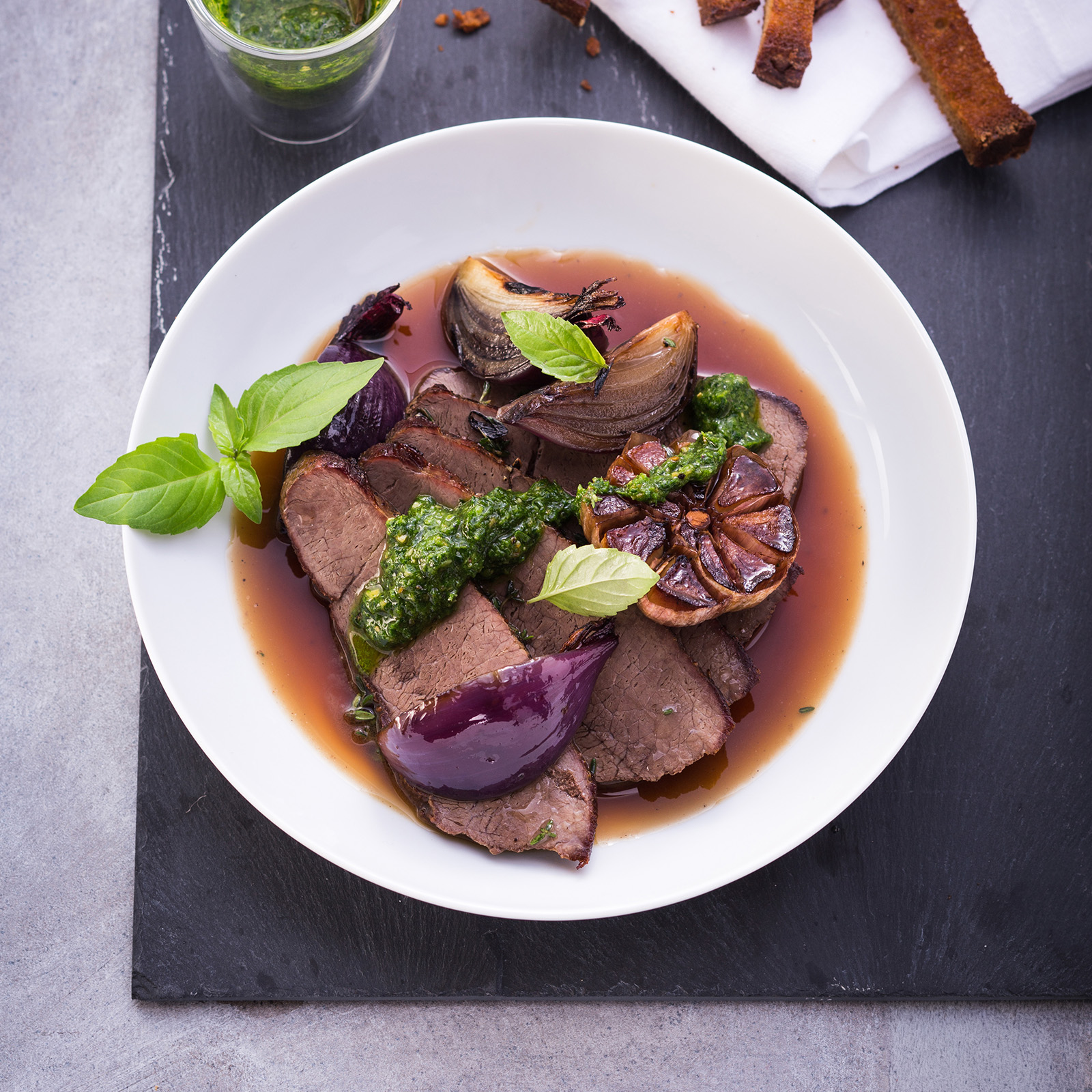 Aufgeschnittener Wildschweinbraten mit Kräuterpesto, geschmorten roten Zwiebeln, gerösteter Knoblauchknolle und frischem Basilikum