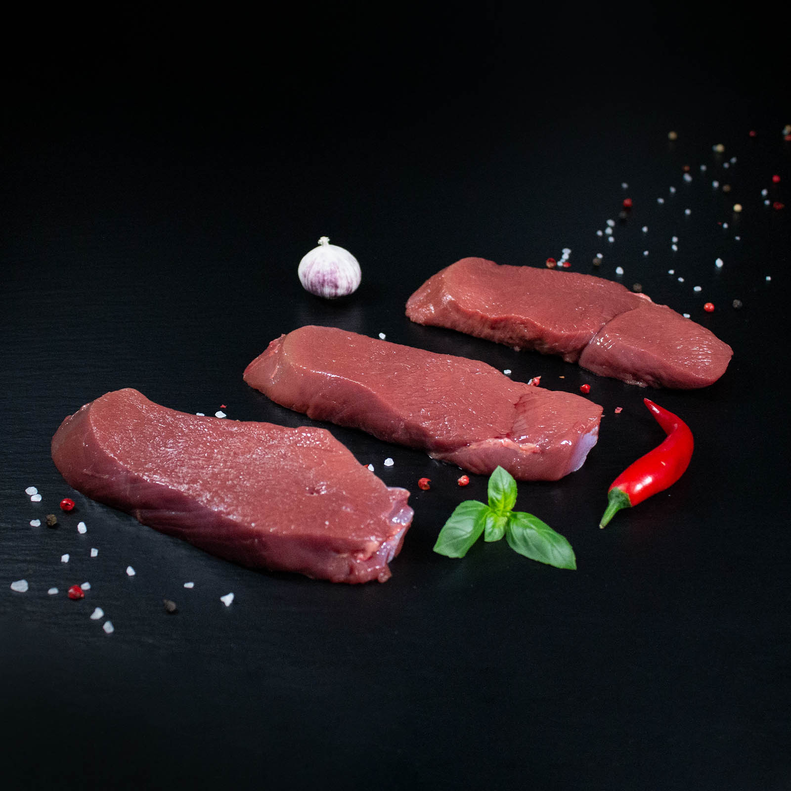 Rohe Hirschsteaks mit Kräutern und Gewürzen auf dunklem Untergrund