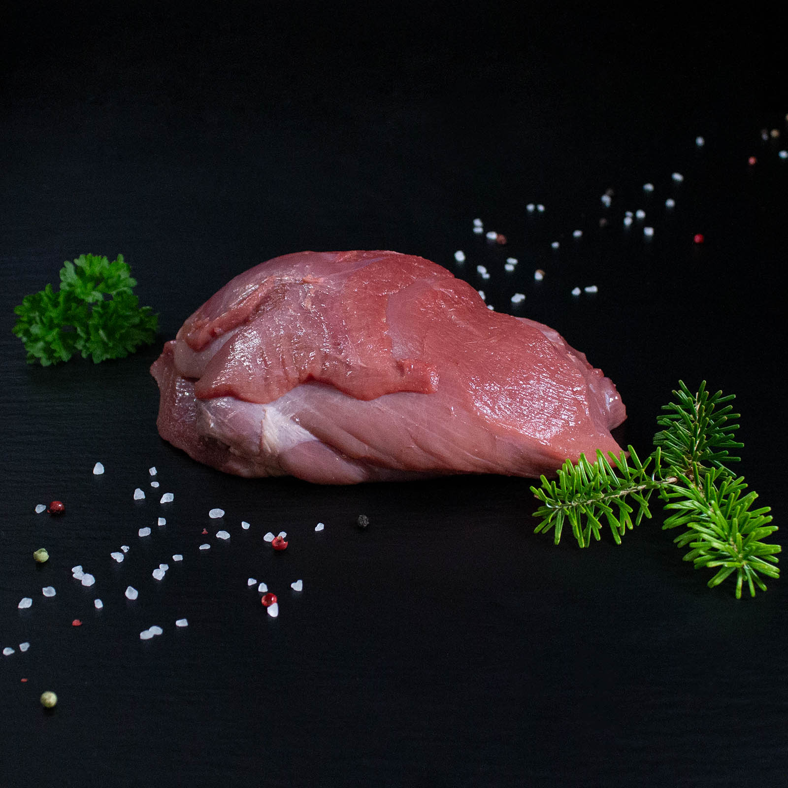 Roher Frischlingsbraten aus der Keule mit Kräutern und Gewürzen auf dunklem Untergrund