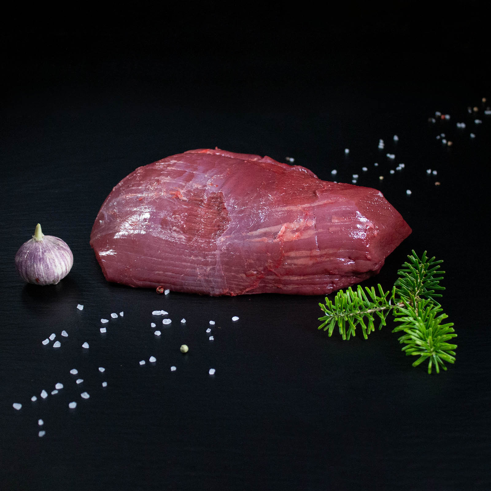 Roher Hirschbraten aus der Keule mit Kräutern und Gewürzen auf dunklem Untergrund
