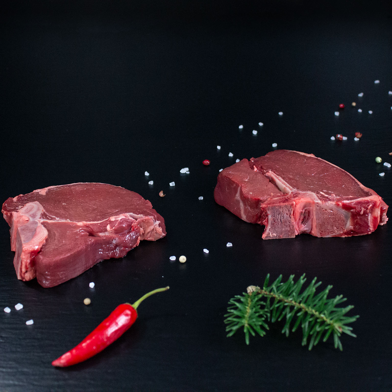 Rohe Hirsch-T-Bone-Steaks mit Kräutern und Gewürzen auf dunklem Untergrund